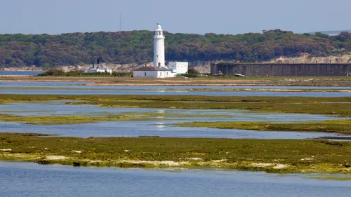 Hurst Point lighthouse This landscape photograph shows Hurst Point Lighthouse situated near Milford-on-Sea, Dorset, United Kingdom, with the location lying close to the Christchurch area. The image was taken in the early afternoon during late spring, featuring the white lighthouse as the central subject, set against a backdrop of green woodland and the historic fortifications of the Hurst Castle. Shallow coastal waters and tidal marshes are visible in the foreground, adding habitat detail typical for this part of the English coast. The clear sky and vibrant greenery suggest the season is spring, and the clarity of the light confirms the time of day as afternoon. The composition highlights the architectural features of Hurst Point Lighthouse amid the natural environment of the Dorset coastline.
